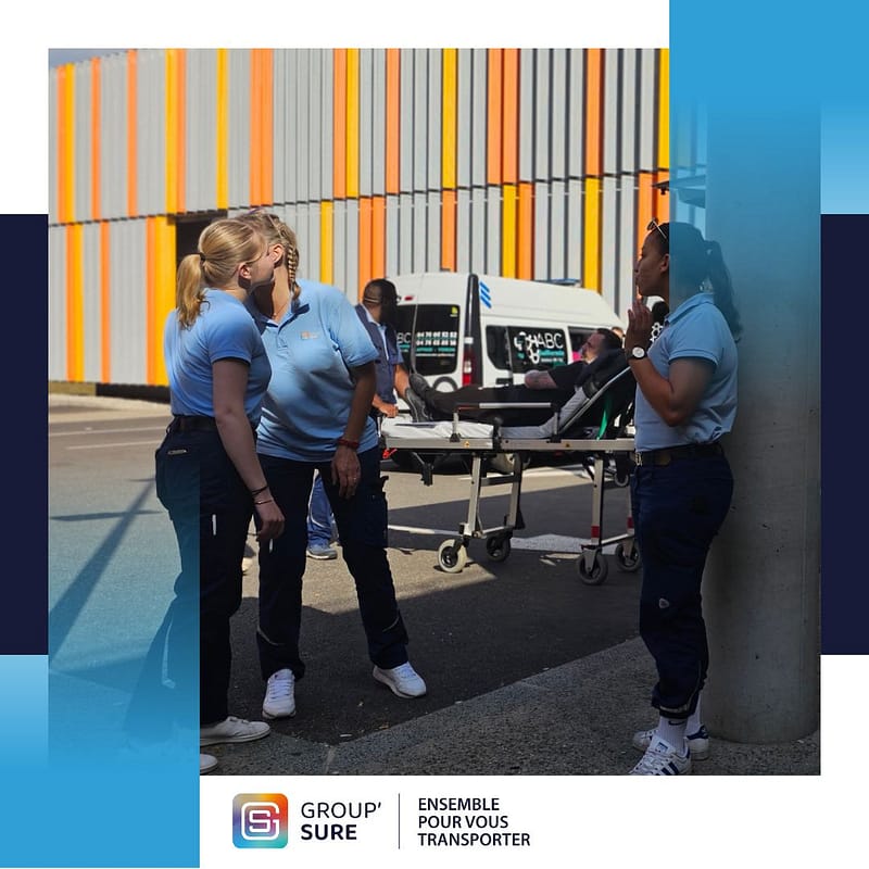 Trois ambulancières de Group’Sure échangent à l’extérieur d’un établissement, avec une ambulance et un brancard en arrière-plan.